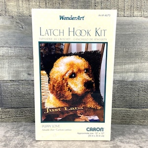 May include: A WonderArt latch hook kit featuring a golden retriever puppy. The kit includes the text "Just Labs" and "Puppy Love". The approximate size is 12" x 12". The box has a green border and is set against a wooden background.