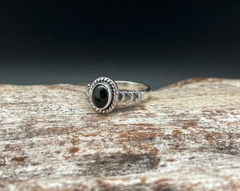 May include: A silver ring featuring a black oval gemstone set in an ornate bezel. The band is decorated with a repeating crescent moon pattern.