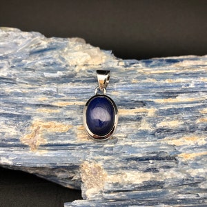 May include: A silver pendant with a dark blue oval gemstone set in a silver bezel. The pendant is on a blue and white background.