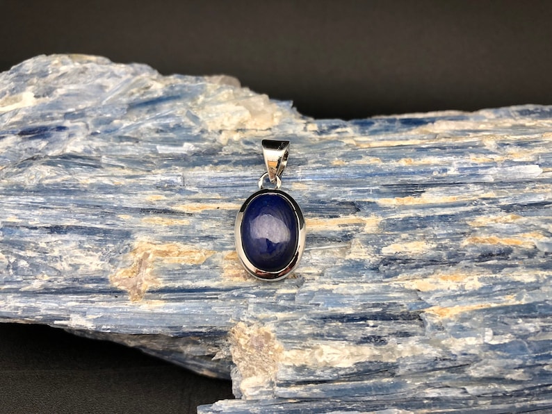 May include: A silver pendant with a dark blue oval gemstone set in a silver bezel. The pendant is on a blue and white background.