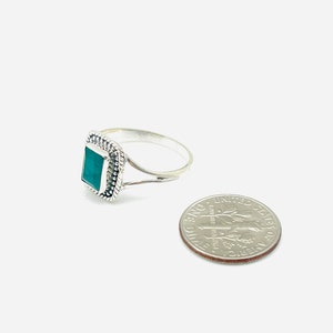 Puede incluir: Un anillo de plata con una piedra verde cuadrada engastada en un borde decorativo. El anillo se muestra junto a una moneda de diez centavos de d&oacute;lar estadounidense para comparar el tama&ntilde;o.