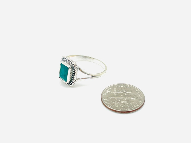 Puede incluir: Un anillo de plata con una piedra verde cuadrada engastada en un borde decorativo. El anillo se muestra junto a una moneda de diez centavos de d&oacute;lar estadounidense para comparar el tama&ntilde;o.
