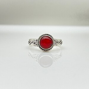 May include: A silver ring with a round, bright red stone. The stone is encircled by a twisted rope design, and the band features decorative swirls on each side. The ring is displayed on a reflective surface.