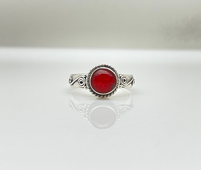 May include: A silver ring with a round, bright red stone. The stone is encircled by a twisted rope design, and the band features decorative swirls on each side. The ring is displayed on a reflective surface.