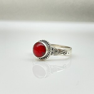 May include: A silver ring featuring a large, round, vibrant red gemstone. The gemstone is set in a decorative bezel with a twisted rope design. The band has an ornate swirl pattern on the side.