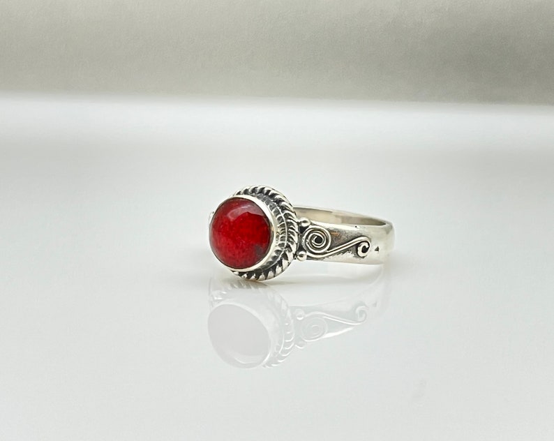 May include: A silver ring featuring a large, round, vibrant red gemstone. The gemstone is set in a decorative bezel with a twisted rope design. The band has an ornate swirl pattern on the side.