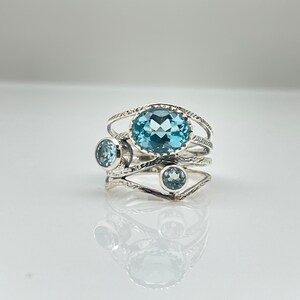 May include: A silver ring with a large oval blue topaz gemstone and two smaller round blue topaz gemstones. The ring has a delicate, intricate design.