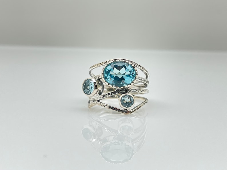 May include: A silver ring with a large oval blue topaz gemstone and two smaller round blue topaz gemstones. The ring has a delicate, intricate design.