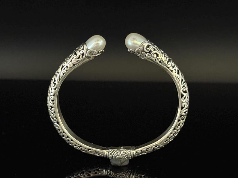 May include: Silver-toned cuff bracelet with ornate, swirling filigree design. The bracelet features a pearl at each end. The open design allows for easy wear. A beautiful piece of jewelry.