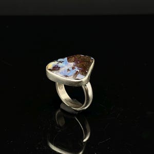 May include: A silver ring featuring a large, irregularly shaped opal stone. The opal displays a mix of colors, including blue, brown, and white. The ring has a simple, polished band and a bezel setting that holds the stone securely.