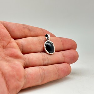 Puede incluir: Colgante de plata con una piedra negra de forma ovalada. El colgante se sostiene en una mano sobre un fondo blanco. La piedra es lisa y pulida, y el engaste de plata tiene un borde texturizado.