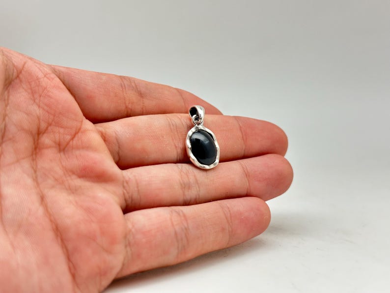 Puede incluir: Colgante de plata con una piedra negra de forma ovalada. El colgante se sostiene en una mano sobre un fondo blanco. La piedra es lisa y pulida, y el engaste de plata tiene un borde texturizado.