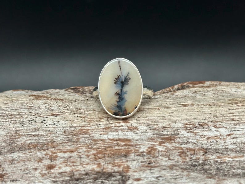 K&ouml;nnte beinhalten: Ovaler Silberring mit einem Dendritachat-Stein. Der Stein zeigt schwarze und blaue Einschl&uuml;sse auf cremewei&szlig;em Grund. Der Ring ist auf einem strukturierten Silberband gefasst. Ein einzigartiges, handgefertigtes Schmuckst&uuml;ck.