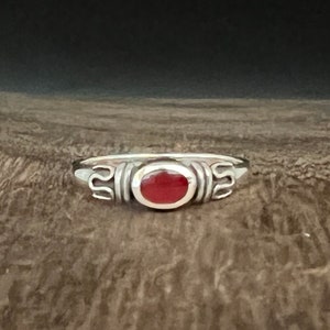 May include: A silver ring with a red oval gemstone set in a bezel setting. The ring has a decorative design on the band.