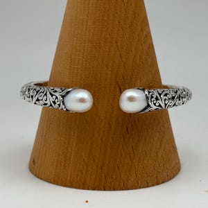 May include: A silver cuff bracelet with an open design, featuring intricate scrollwork and two large, round, white pearls at the ends. The bracelet is displayed on a wooden stand.