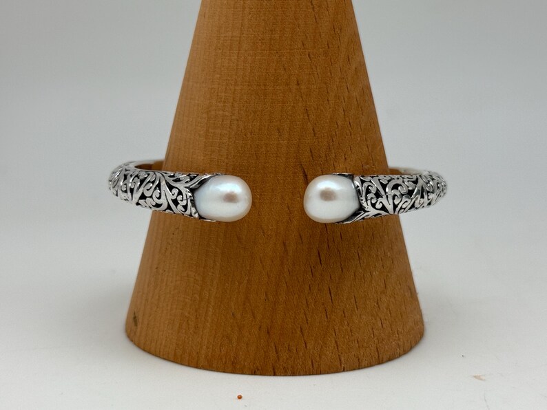 May include: A silver cuff bracelet with an open design, featuring intricate scrollwork and two large, round, white pearls at the ends. The bracelet is displayed on a wooden stand.