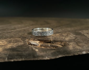 May include: A silver ring with an engraved leaf and branch pattern. The ring is set against a dark wood surface. The ring's design features detailed botanical elements, creating a nature-inspired aesthetic. The ring is a band style.