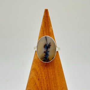 K&ouml;nnte beinhalten: Ein Silberring mit einem ovalen Dendritachat-Stein. Der Stein hat eine cremefarbene Basis mit dunkelblauen und schwarzen, farnartigen Einschl&uuml;ssen. Der Ring wird auf einem Holzkegel pr&auml;sentiert.