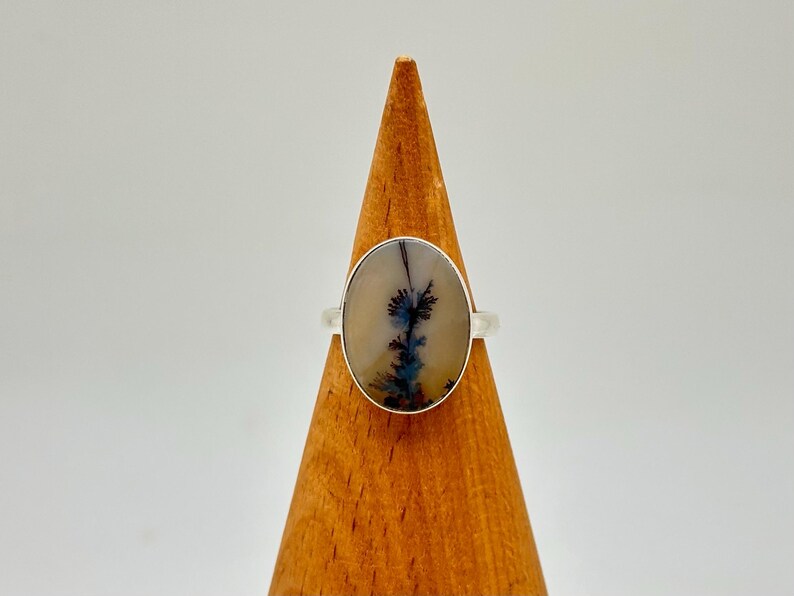 K&ouml;nnte beinhalten: Ein Silberring mit einem ovalen Dendritachat-Stein. Der Stein hat eine cremefarbene Basis mit dunkelblauen und schwarzen, farnartigen Einschl&uuml;ssen. Der Ring wird auf einem Holzkegel pr&auml;sentiert.