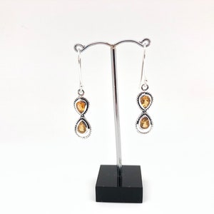 May include: A pair of silver drop earrings featuring two stacked, pear-shaped yellow citrine gemstones in each earring. The stones are set in twisted silver metal frames, suspended from silver hook wires on a metal display stand.