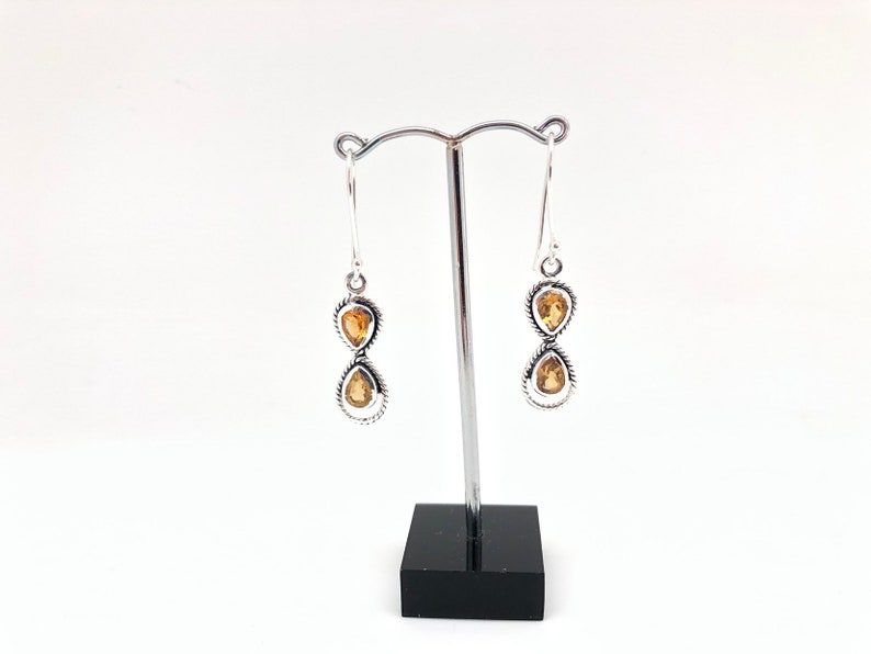 May include: A pair of silver drop earrings featuring two stacked, pear-shaped yellow citrine gemstones in each earring. The stones are set in twisted silver metal frames, suspended from silver hook wires on a metal display stand.