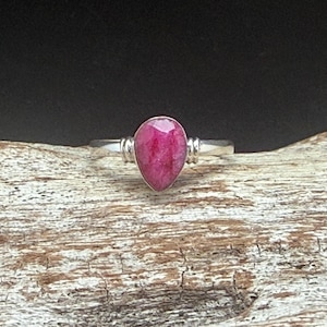 Puede incluir: Anillo de plata con una gema de rubí en forma de lágrima. El rubí es de un color rosa intenso, engastado en una banda sencilla. El anillo se muestra sobre una superficie de madera texturizada sobre un fondo oscuro.