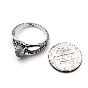 May include: A silver ring featuring a moonstone set within a twisted branch design. The ring is shown alongside a UK ten pence coin for scale.