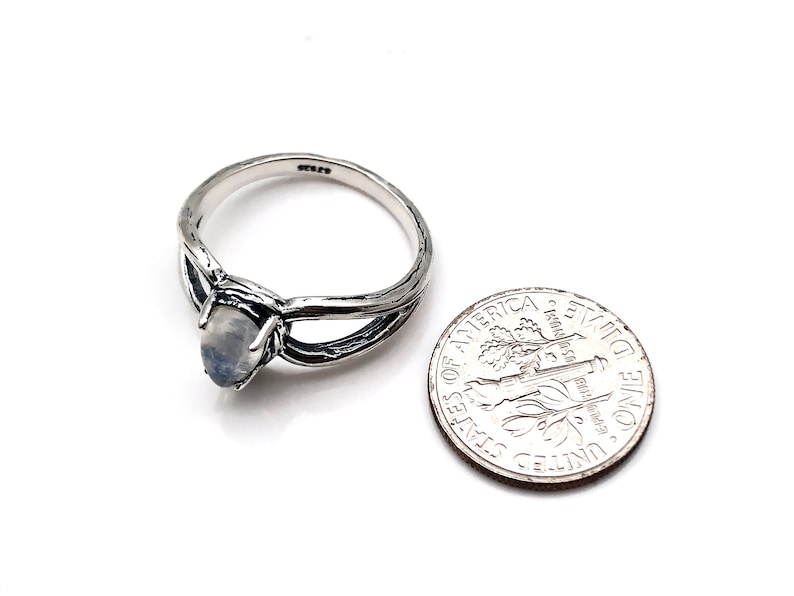 May include: A silver ring featuring a moonstone set within a twisted branch design. The ring is shown alongside a UK ten pence coin for scale.