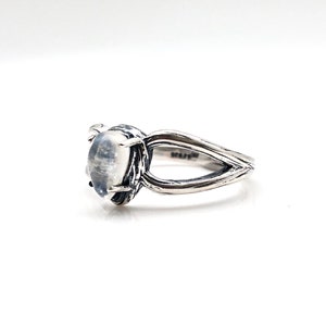 May include: A silver ring featuring a white moonstone set in a textured band. The band is shaped like a branch with two prongs.