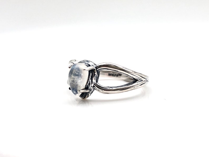 May include: A silver ring featuring a white moonstone set in a textured band. The band is shaped like a branch with two prongs.