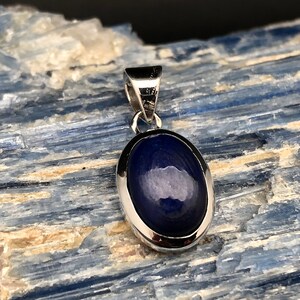 May include: A silver pendant with a dark blue oval gemstone. The pendant is set in a silver bezel and has a bail for attaching a chain.