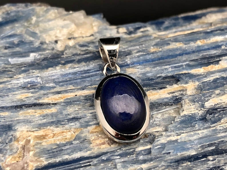 May include: A silver pendant with a dark blue oval gemstone. The pendant is set in a silver bezel and has a bail for attaching a chain.