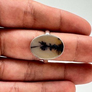 K&ouml;nnte beinhalten: Ovaler Ring mit einem Dendritachat-Stein und einem Silberband. Der Achat hat eine cremefarbene Basis mit schwarzen, baumartigen Einschl&uuml;ssen. Der Ring ist auf einem Silberband gefasst und zeigt ein von der Natur inspiriertes Design.