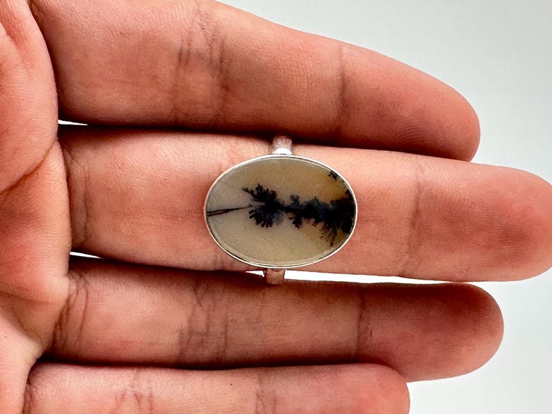 K&ouml;nnte beinhalten: Ovaler Ring mit einem Dendritachat-Stein und einem Silberband. Der Achat hat eine cremefarbene Basis mit schwarzen, baumartigen Einschl&uuml;ssen. Der Ring ist auf einem Silberband gefasst und zeigt ein von der Natur inspiriertes Design.