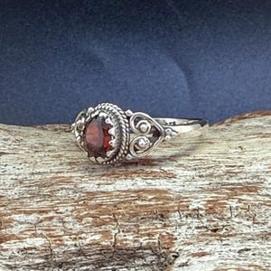 May include: A silver ring featuring a central round, deep red gemstone set within a twisted rope border. The band displays intricate metalwork with heart-shaped filigree details on the sides, creating a decorative and detailed appearance.