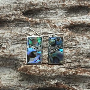 May include: A pair of rectangular silver earrings with iridescent abalone shell inlays. The earrings feature a decorative silver filigree design and a hook closure. The abalone shell displays shades of blue, green, and purple.