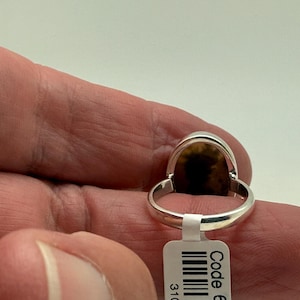 Pu&ograve; includere: Un anello d'argento con una pietra ovale marrone e nera. L'anello &egrave; indossato su un dito e ha un'etichetta con codice a barre con il testo "Code 6".