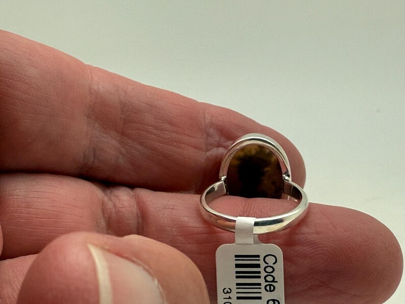 Pu&ograve; includere: Un anello d'argento con una pietra ovale marrone e nera. L'anello &egrave; indossato su un dito e ha un'etichetta con codice a barre con il testo "Code 6".