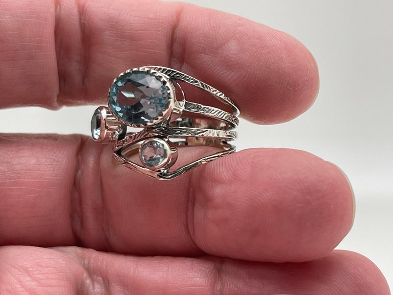 May include: A silver ring with three blue topaz gemstones. The ring has a delicate, intricate design with multiple bands.