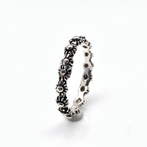 May include: A silver-colored ring featuring a repeating floral pattern. The ring has a detailed design with small, daisy-like flowers around the band. The ring is set against a white background.
