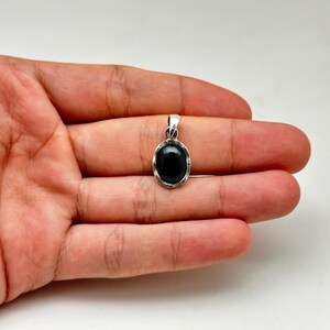 Puede incluir: Un colgante de plata con una piedra preciosa negra ovalada. El colgante se sostiene en una mano sobre un fondo blanco. La piedra preciosa es lisa y pulida, y el engaste de plata tiene un dise&ntilde;o sencillo.