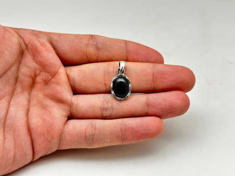 Puede incluir: Un colgante de plata con una piedra preciosa negra ovalada. El colgante se sostiene en una mano sobre un fondo blanco. La piedra preciosa es lisa y pulida, y el engaste de plata tiene un dise&ntilde;o sencillo.