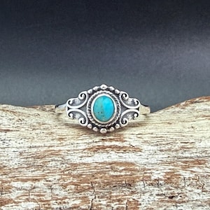 May include: A silver ring with an oval turquoise stone at the center. The ring has an ornate design with swirls and beaded accents around the stone. The ring is set on a wooden surface.