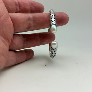 May include: A silver-colored cuff bracelet with an open design, featuring intricate scrollwork and two large, white, teardrop-shaped pearls. The bracelet is displayed against a white background.