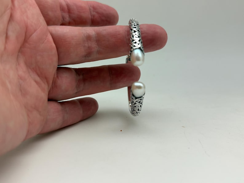 May include: A silver-colored cuff bracelet with an open design, featuring intricate scrollwork and two large, white, teardrop-shaped pearls. The bracelet is displayed against a white background.