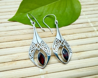 May include: A pair of silver drop earrings featuring a marquise-cut deep red garnet gemstone suspended within a teardrop-shaped frame. The metal frame displays an intricate openwork scroll pattern, and each earring is attached to a curved hook.