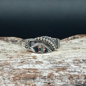 May include: A silver ring with an ornate design. The ring features a central rectangular element with a detailed pattern and is surrounded by small, raised dots. The ring is set against a wooden surface.