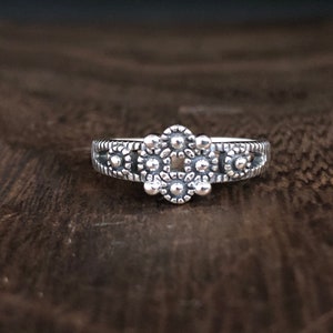May include: A silver ring with a detailed design featuring a central cluster of five round, raised circles surrounded by smaller circles. The ring band is also decorated with small circles.