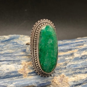 May include: A silver ring with a large, oval-shaped green gemstone set in a detailed silver bezel. The ring is on a blue and white background.