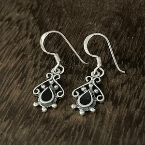 May include: A pair of silver dangle earrings with black teardrop-shaped stones. The earrings have a delicate, ornate design.